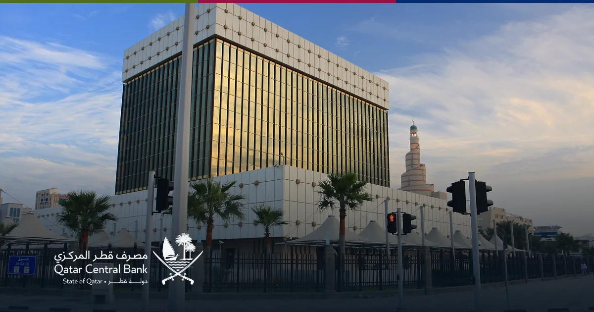 مبنى مصرف قطر المركزي في الدوحة عند الغروب، وخلفه مأذنة مسجد الفنار الحلزونية، مع شعار المصرف في أسفل الصورة